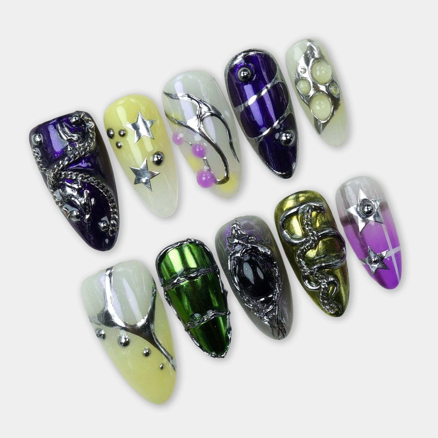 A set of eclectic press-on nails featuring chrome green, purple, yellow, and clear bases with metallic stars, chains, beads, and 3D art details.