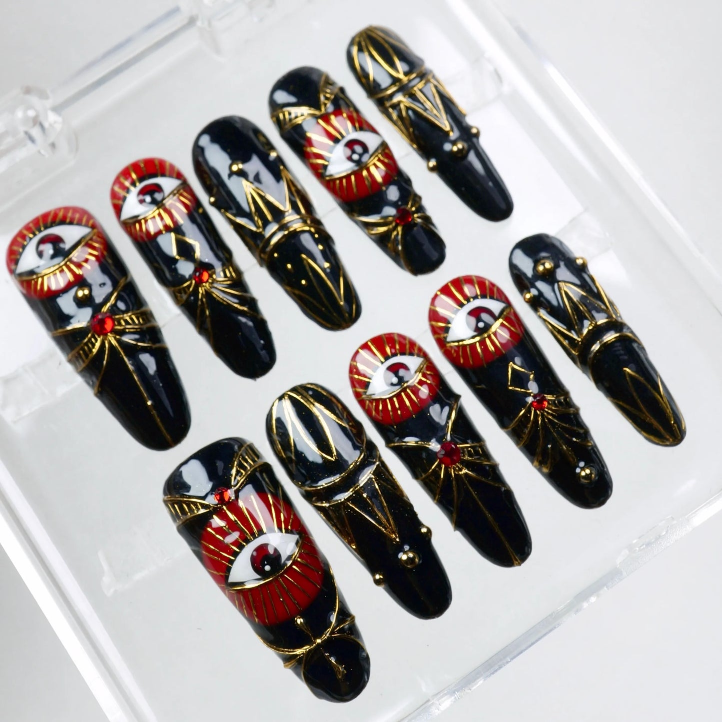 Press-on nails featuring intricate red and gold evil eyes, royal black base, and geometric gold foil elements from Donailsart