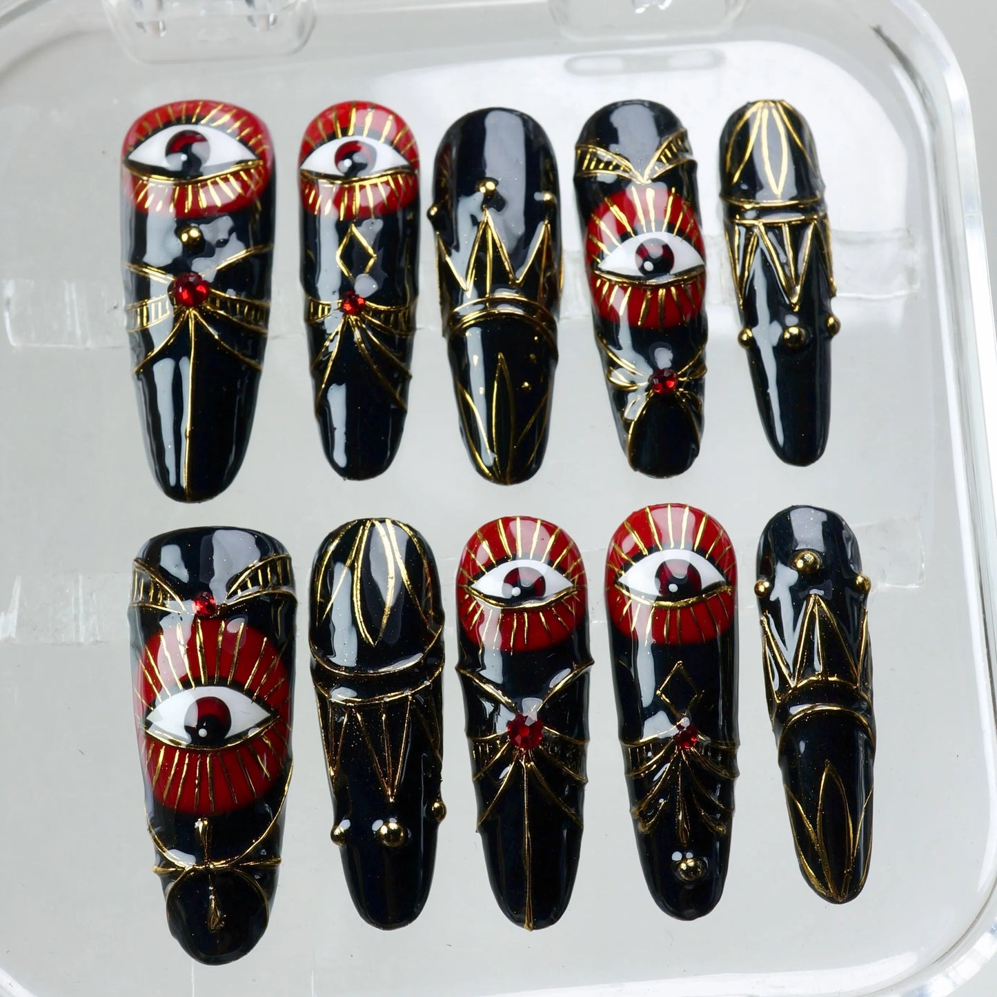 Dramatic press-on nails inspired by ancient symbolism, featuring bold red eyes, deep black polish, and golden geometry from Donailsart