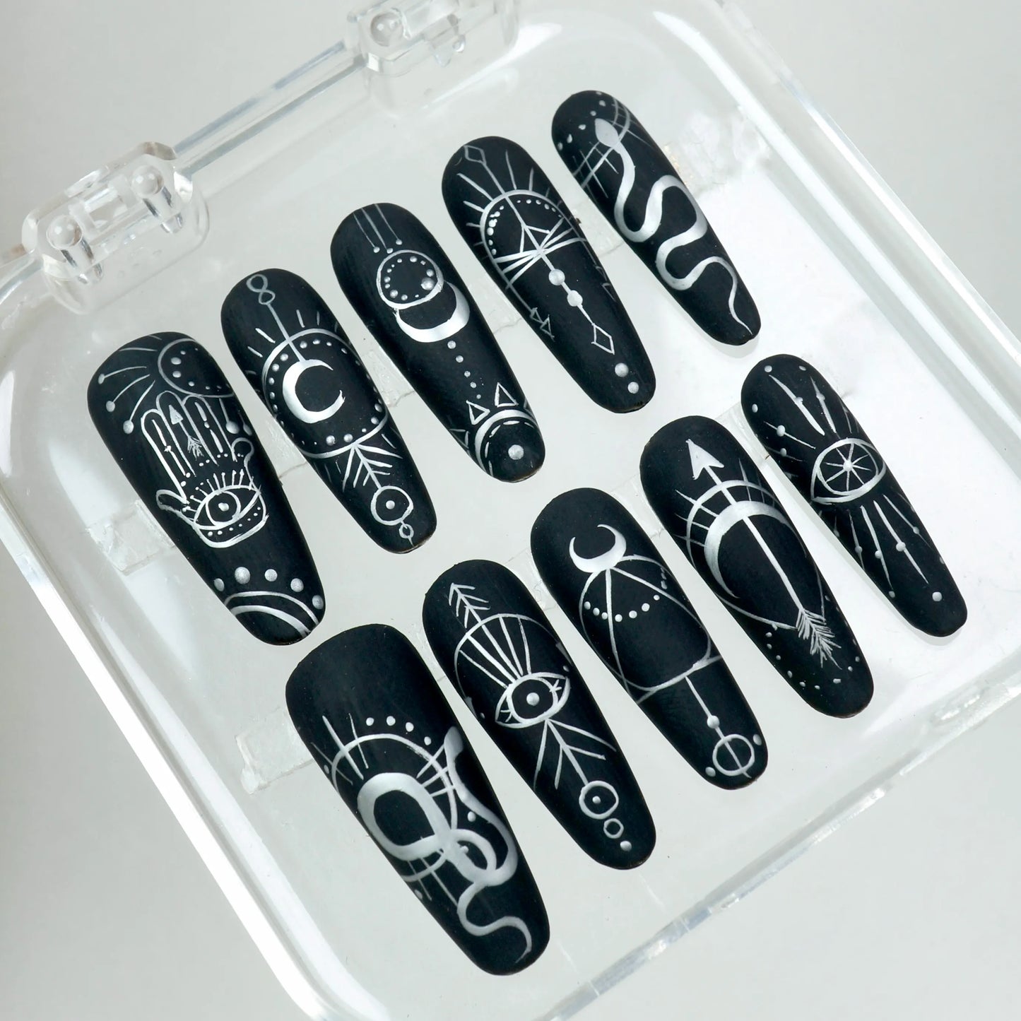 Matte black press-on nails featuring white hand-drawn mystical motifs like crescent moons, evil eyes, and geometric lines