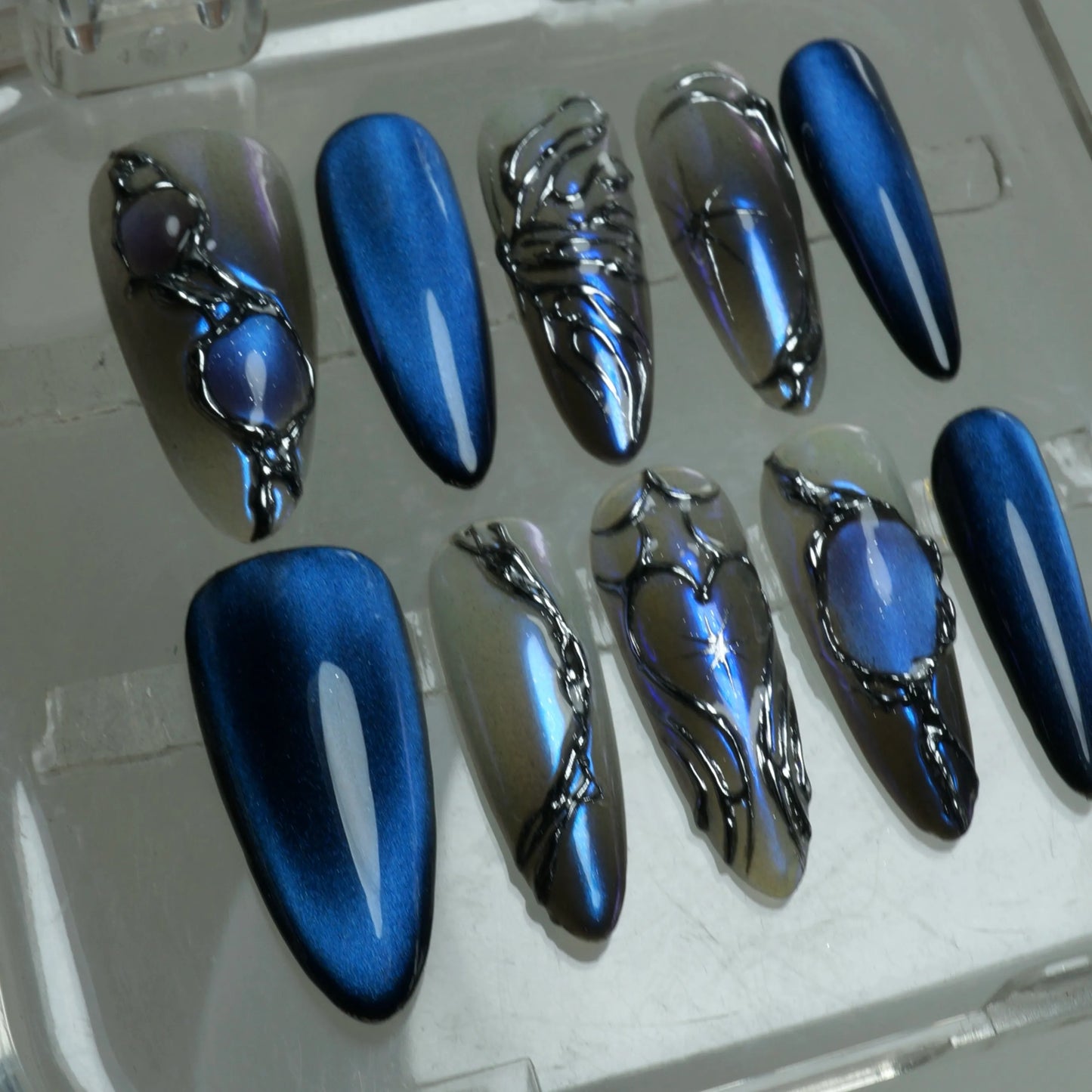 Press-on nails designed with liquid chrome effects, deep sapphire hues, and moody fantasy-inspired accents