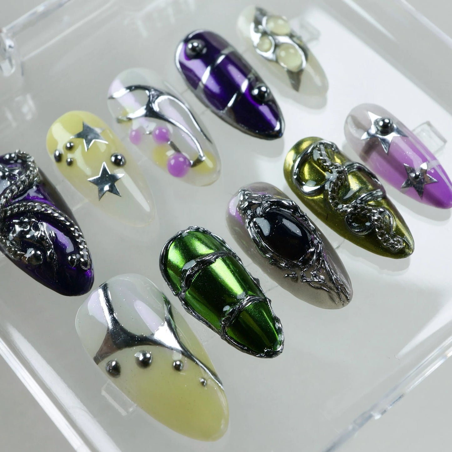 A set of eclectic press-on nails featuring chrome green, purple, yellow, and clear bases with metallic stars, chains, beads, and 3D art details.