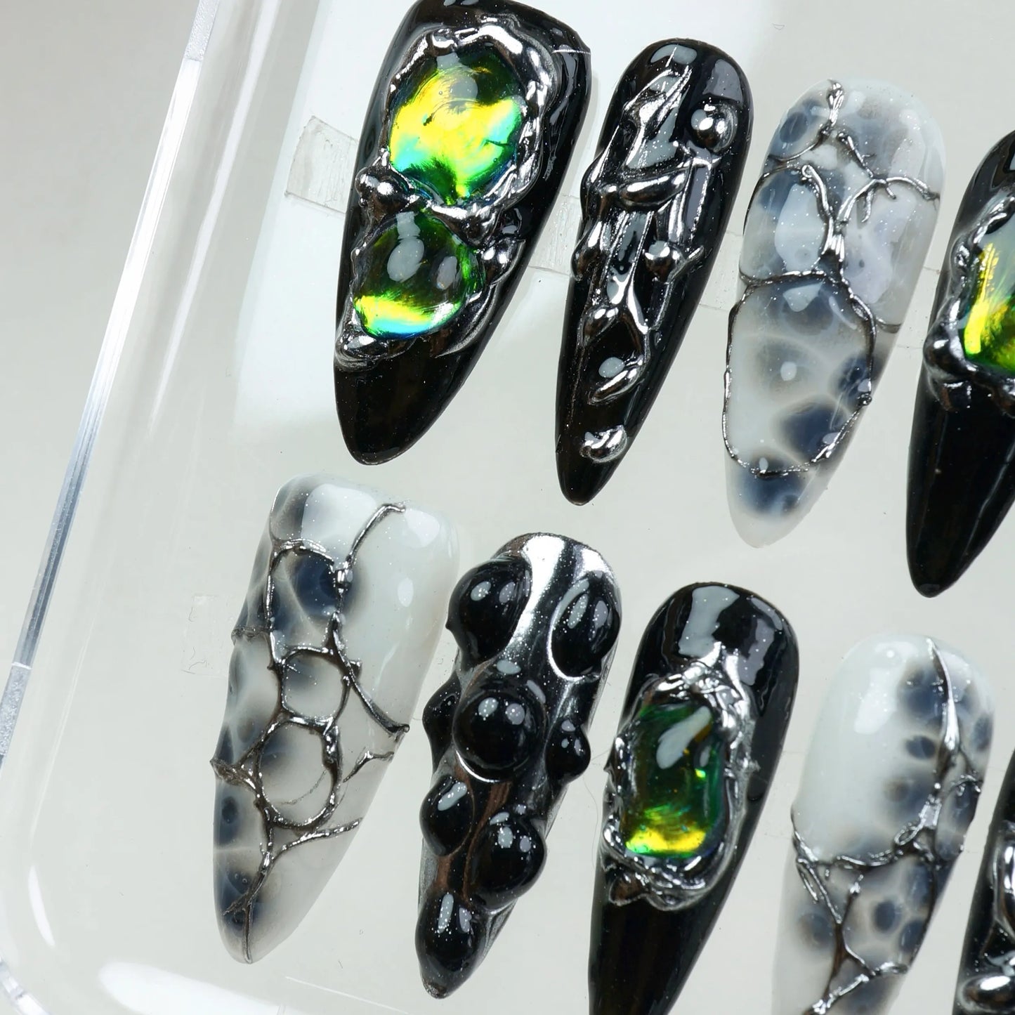 Black and grey press-on nails with 3D chrome textures, glass-like droplets, and holographic gemstone details.