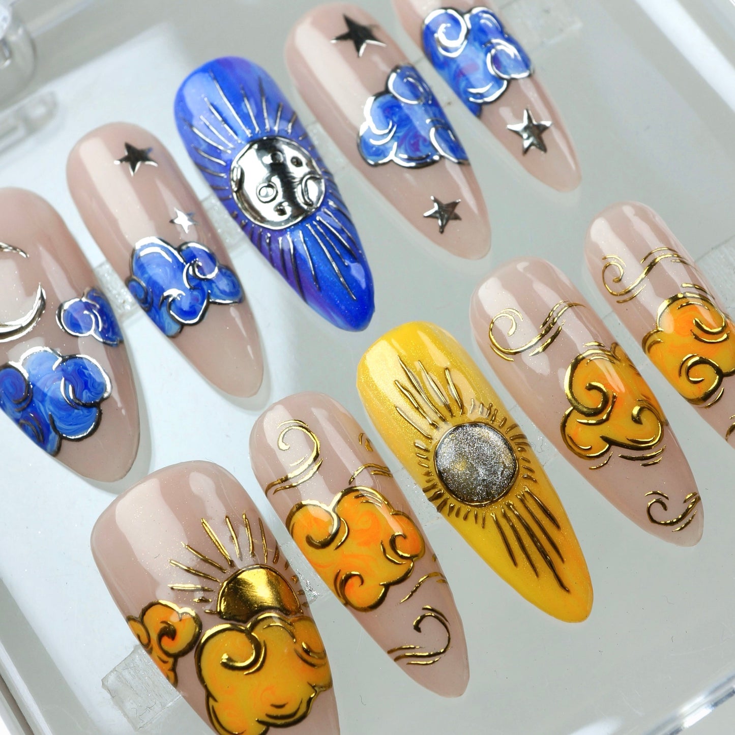 Day & Night press-on nails macro close-up in clear case – night side showing blue cloud nails with silver crescent and sun-face charm; day side showing orange cloud nails with gold sun rays and glitter sun charm on yellow accent nail