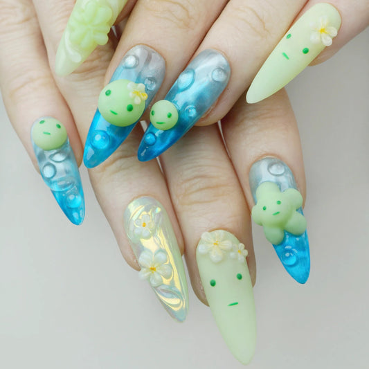 Close-up of Smiski Glow in the Dark stiletto press-on nails worn on hand, aqua blue aurora nails with bubble art and yellow-green nails featuring 3D Smiski figurine charm and white plumeria flowers