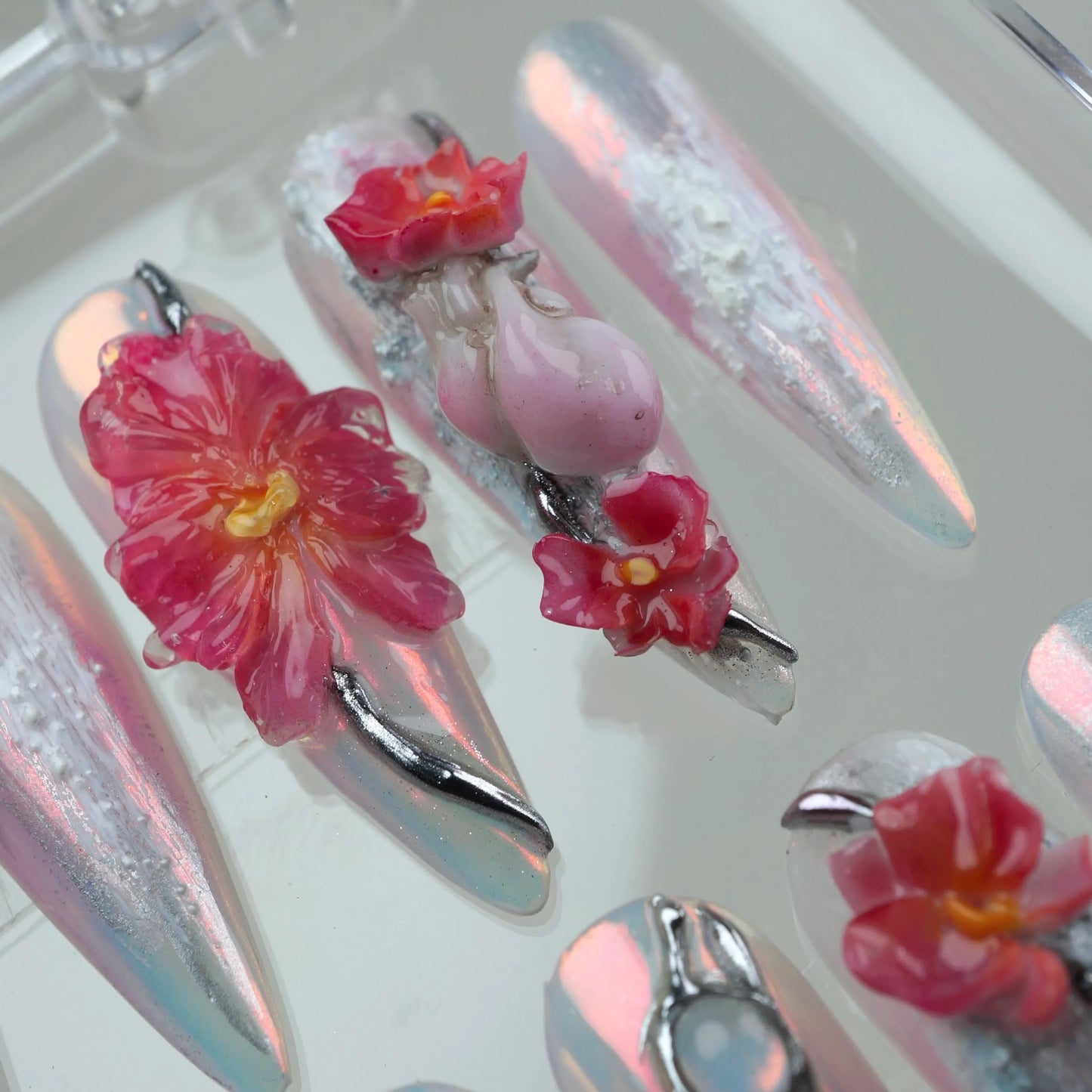 Extreme close-up of Tropical Requiem press-on nail art featuring oversized 3D red hibiscus flower, pink rose bud charm, red orchid, and silver fang charms on iridescent aurora white stiletto nails