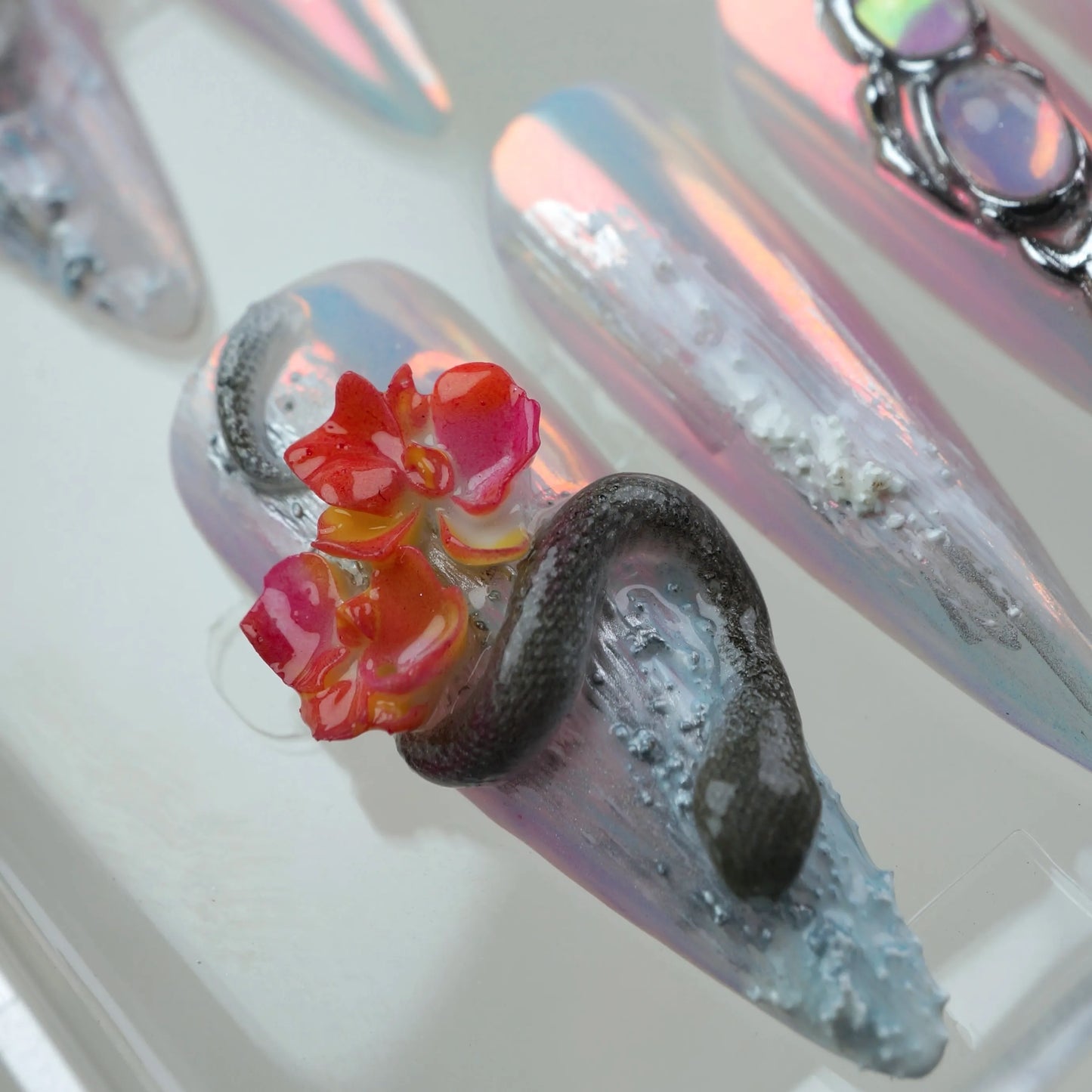 Iridescent summer nails art with floral accents, snake and skull sculptures, and icy chrome finish