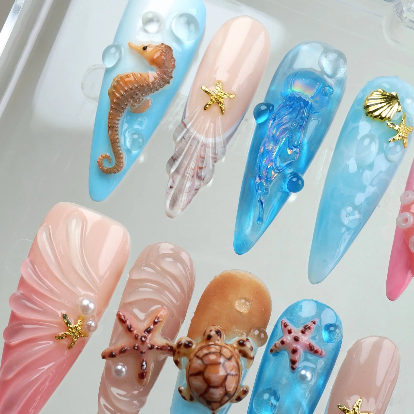 Aquatic press-on nail set viewed from above showing seahorse, iridescent jellyfish, shell, fan shell, sea turtle, and starfish stiletto nails in baby blue, blush pink, sandy beige, and aqua blue with pearl and bubble water art