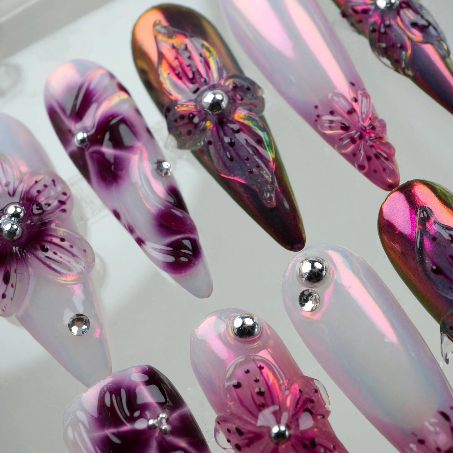 Close-up detail of Velvet Petals press-on nails in acrylic case, featuring 3D translucent purple-pink flower charms, frosted white stiletto nails with purple ink floral swirls, and dark iridescent chrome nails with silver bead details