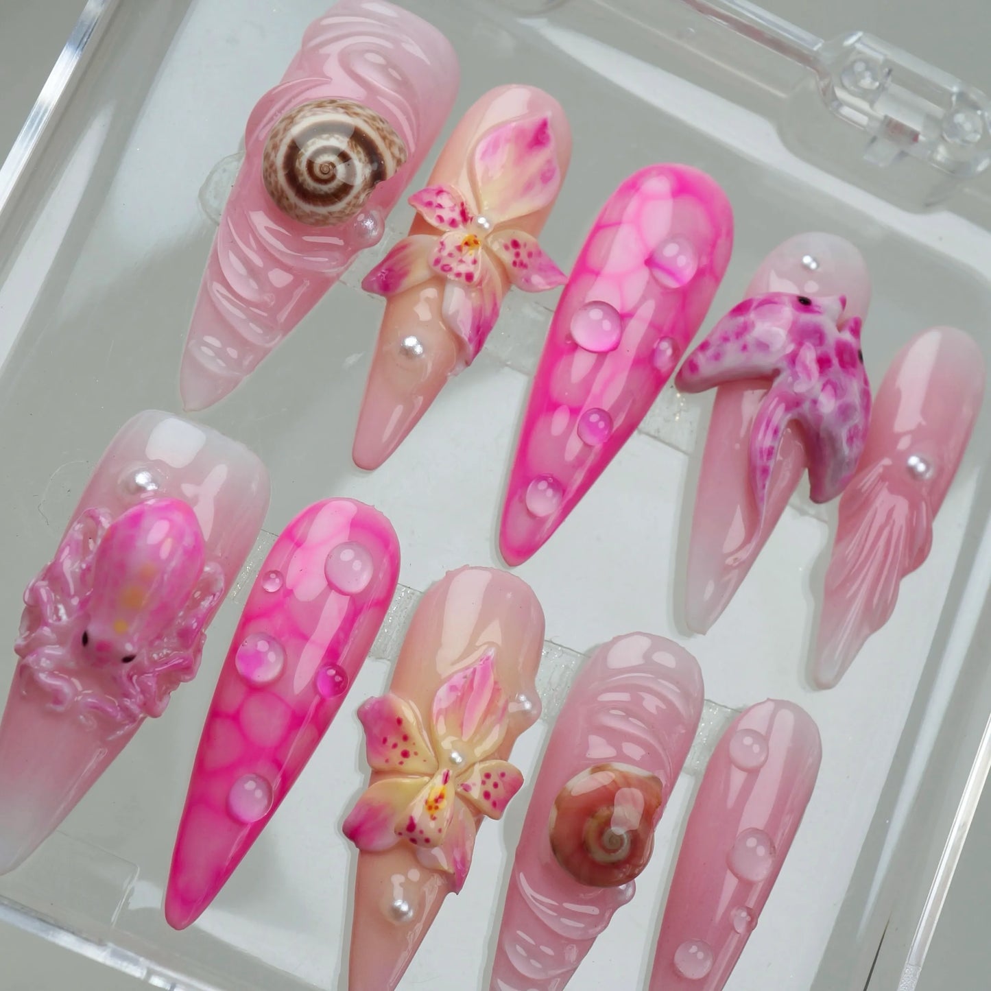 Rosy Tide press-on nail set top-down view in clear acrylic case, showcasing 10 stiletto nails with pink ocean wave swirl, hot pink bubble art, 3D seashell charms, 3D orchid flower charms, 3D spotted starfish charm, and 3D pink octopus charm