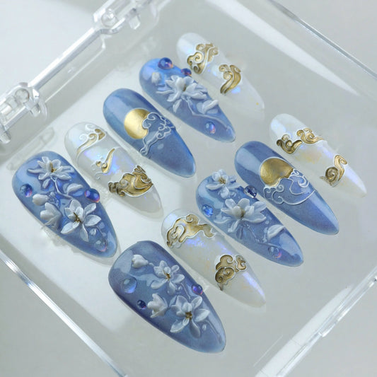 Press-on nails with hand-sculpted white florals, golden moon and cloud details, and soft blue and pearl shimmer base by Donailsart