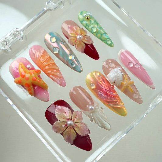 Handcrafted ocean-themed press-on nails with 3D starfish, seashells, and floral designs in pink and iridescent shades, displayed in a clear case