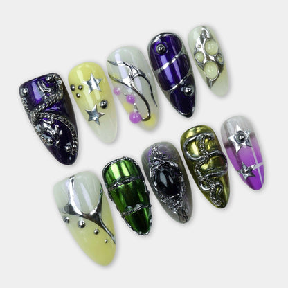 A set of eclectic press-on nails featuring chrome green, purple, yellow, and clear bases with metallic stars, chains, beads, and 3D art details.