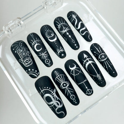Matte black press-on nails featuring white hand-drawn mystical motifs like crescent moons, evil eyes, and geometric lines