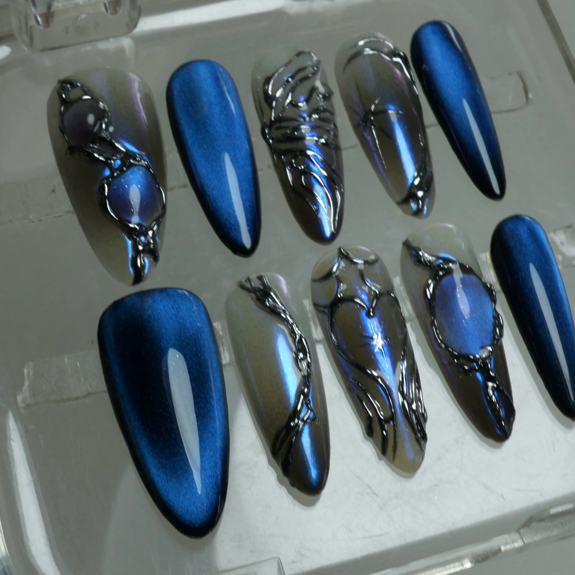 Press-on nails designed with liquid chrome effects, deep sapphire hues, and moody fantasy-inspired accents