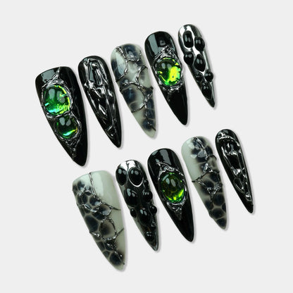 Emerald Enchantment press-on nails – almond-shaped set with black nails featuring silver liquid metal drip art and large iridescent green gemstone, black-and-white marble nails with silver chrome veining, flat lay on white background