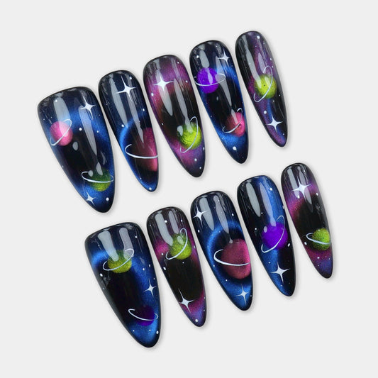 Galaxy-themed press-on nails with shimmering planets, glowing rings, and hand-painted stars on a black and blue space background
