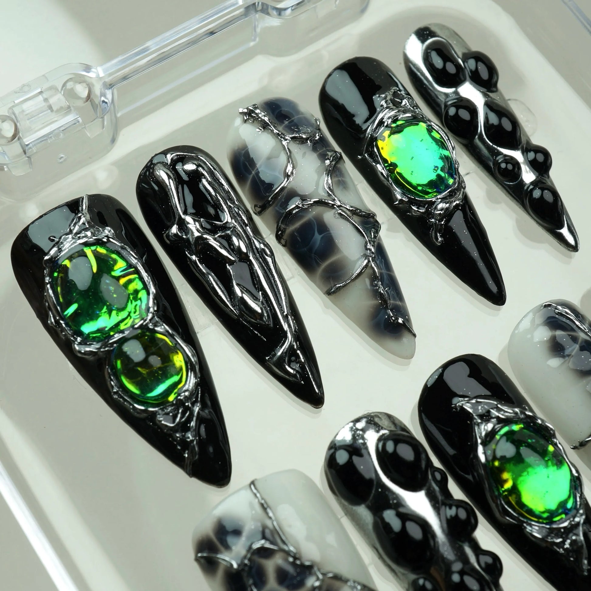 Emerald Enchantment press-on nails close-up in clear acrylic case – detailed view of large iridescent green emerald gemstones and silver chrome liquid metal drip art on black almond nails