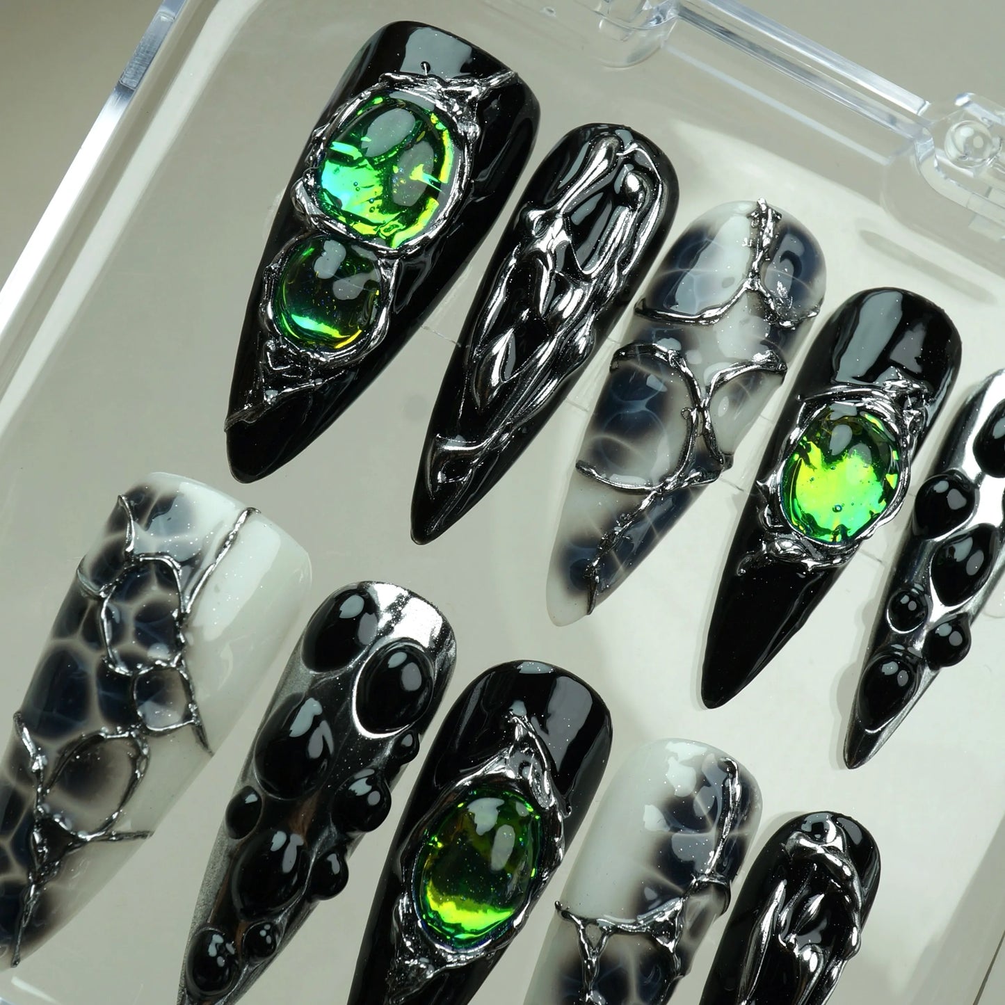Emerald Enchantment press-on nails top-down close-up in clear case – almond nails combining iridescent green gems with silver chrome wire art, black bubble dot accents, and smoky marble ink swirl nails