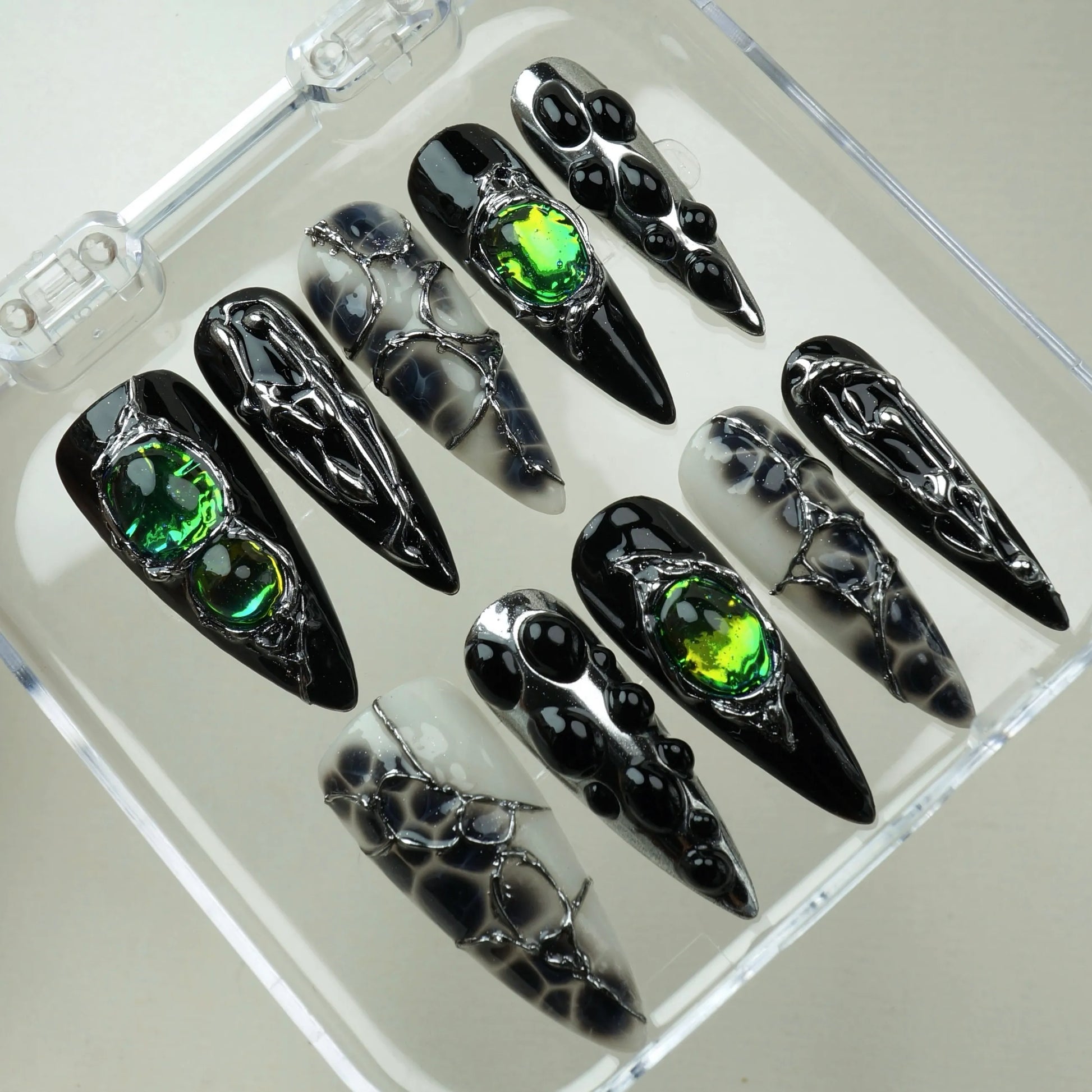 Emerald Enchantment press-on nails in open clear acrylic case – overhead angled view of full mixed design set with silver chrome drip art, iridescent green gemstones, and black-and-white marble ink nails