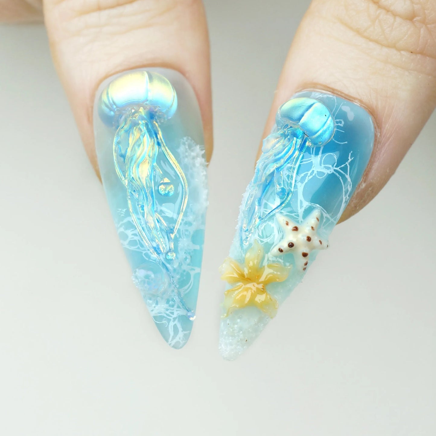 Ocean Bloom press-on nail close-up showing 3D iridescent jellyfish with flowing translucent tentacles and 3D white spotted starfish with orange flower charm on aqua base