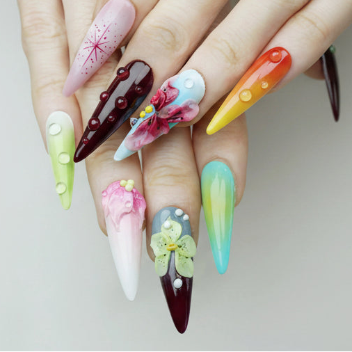 Nymph's Garden stiletto press-on nails worn on hand showing rainbow of colors including pink starburst nail art, dark maroon nail with red bubble dots, baby blue nail with 3D pink orchid and yellow dot accents, and orange-yellow gradient nail