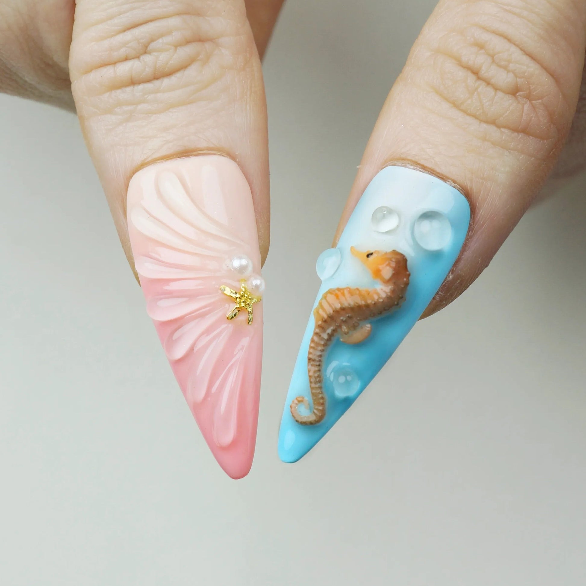 Close-up of Aquatic press-on nails showing blush pink fan shell sculpted nail with pearl and gold starfish charm beside baby blue nail with detailed 3D brown seahorse and pearl water bubble accents