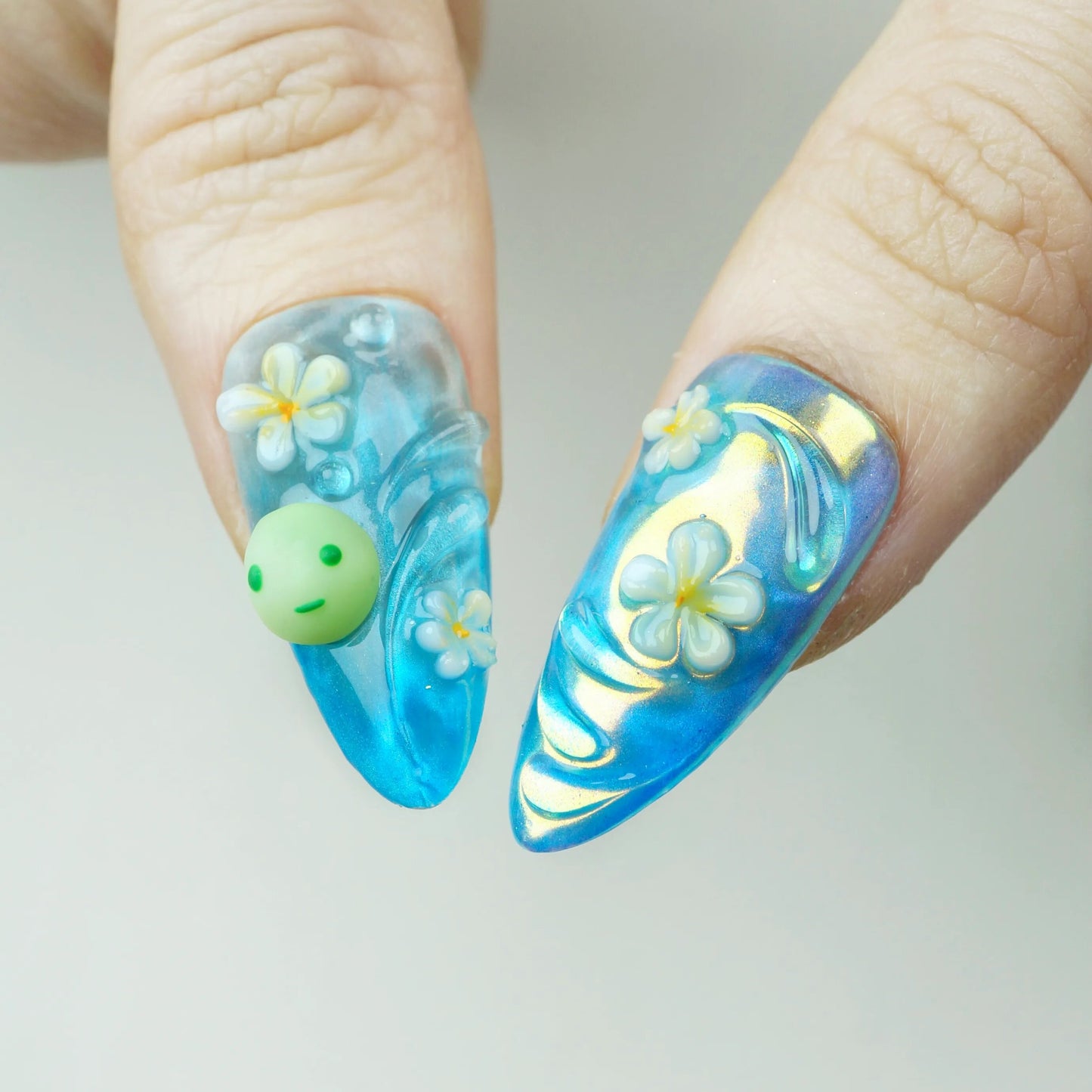 Close-up detail of Smiski Glow in the Dark press-on nails showing aqua blue stiletto nails with 3D Smiski charm, white plumeria flowers, and iridescent aurora finish