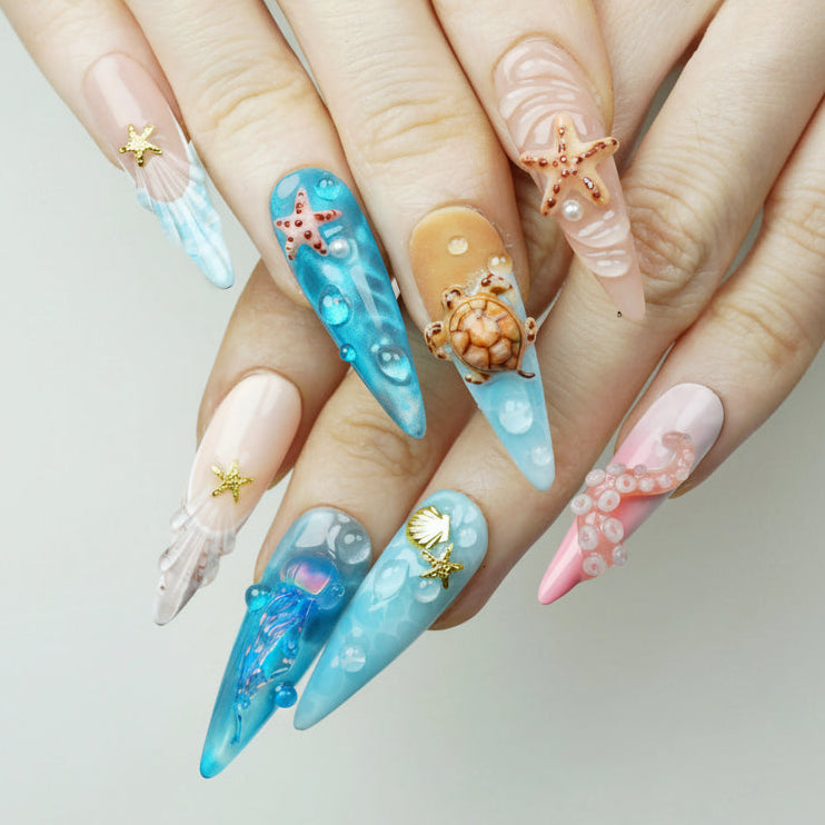 Aquatic press-on nails worn on both hands showing ocean-themed stiletto nail art including 3D sea turtle charm, iridescent jellyfish, starfish, shell fan texture, and bubble water effects on pink, blue, and nude bases
