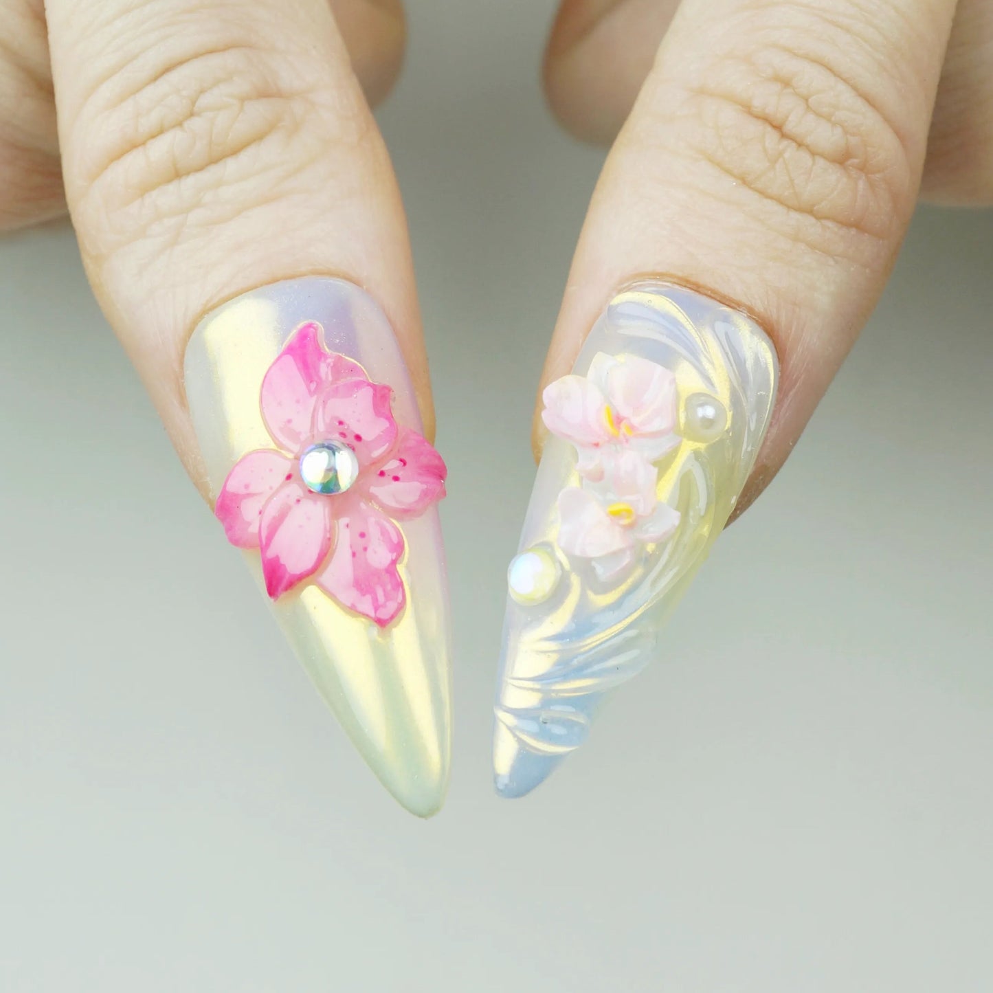 Flower Mirage press-on nail close-up showing 3D vibrant pink orchid flower with AB rhinestone center on gold iridescent nail and soft 3D cherry blossom cluster with pearl on aurora nail