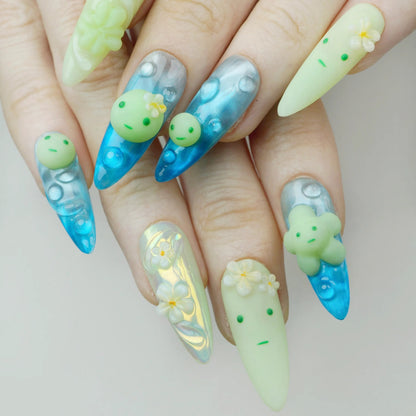 Close-up of Smiski Glow in the Dark stiletto press-on nails worn on hand, aqua blue aurora nails with bubble art and yellow-green nails featuring 3D Smiski figurine charm and white plumeria flowers
