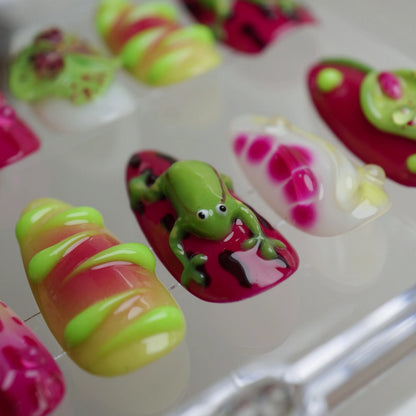 Colorful nail art with fruit designs and a frog figure on a white background