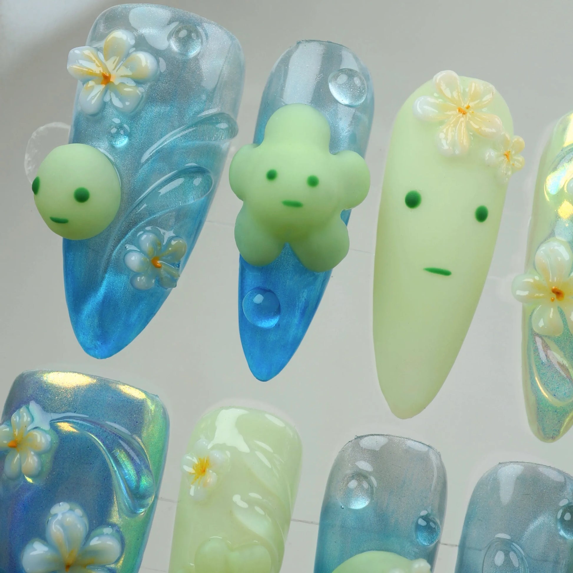 Whimsical smiski nails with pastel green figures, glossy blue gradients, and small 3D flowers for a fun summer vibe