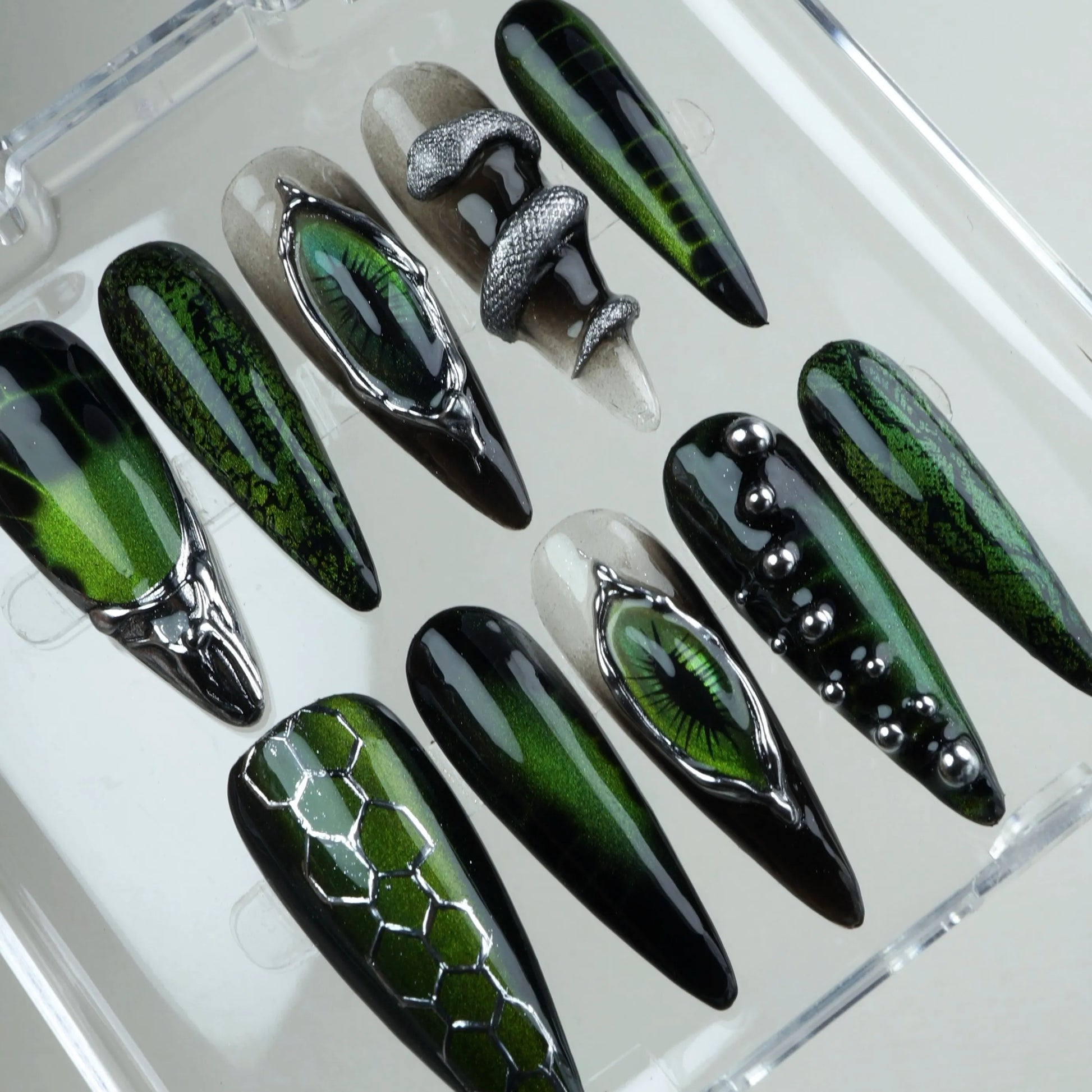 Cat-eye nails design with holographic green gel, silver 3D embellishments, and sci-fi serpent motifs