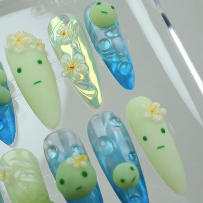 Creative smiski nails with holographic blue bases, smiling green faces, and plumeria-style blossoms