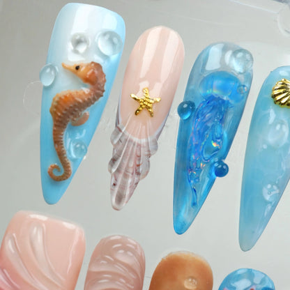 Close-up of Aquatic press-on nails in display case showing baby blue nail with 3D brown seahorse and pearl bubbles, translucent shell jelly nail with gold starfish charm, and blue holographic iridescent jellyfish nail