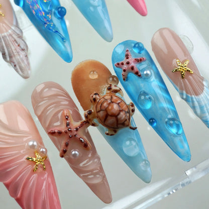 Close-up of Aquatic press-on nails showing blush pink fan shell nail with pearl and gold starfish, sandy beige nail with 3D coral starfish and pearl, amber nail with realistic 3D sea turtle and starfish, and blue water bubble nail with starfish