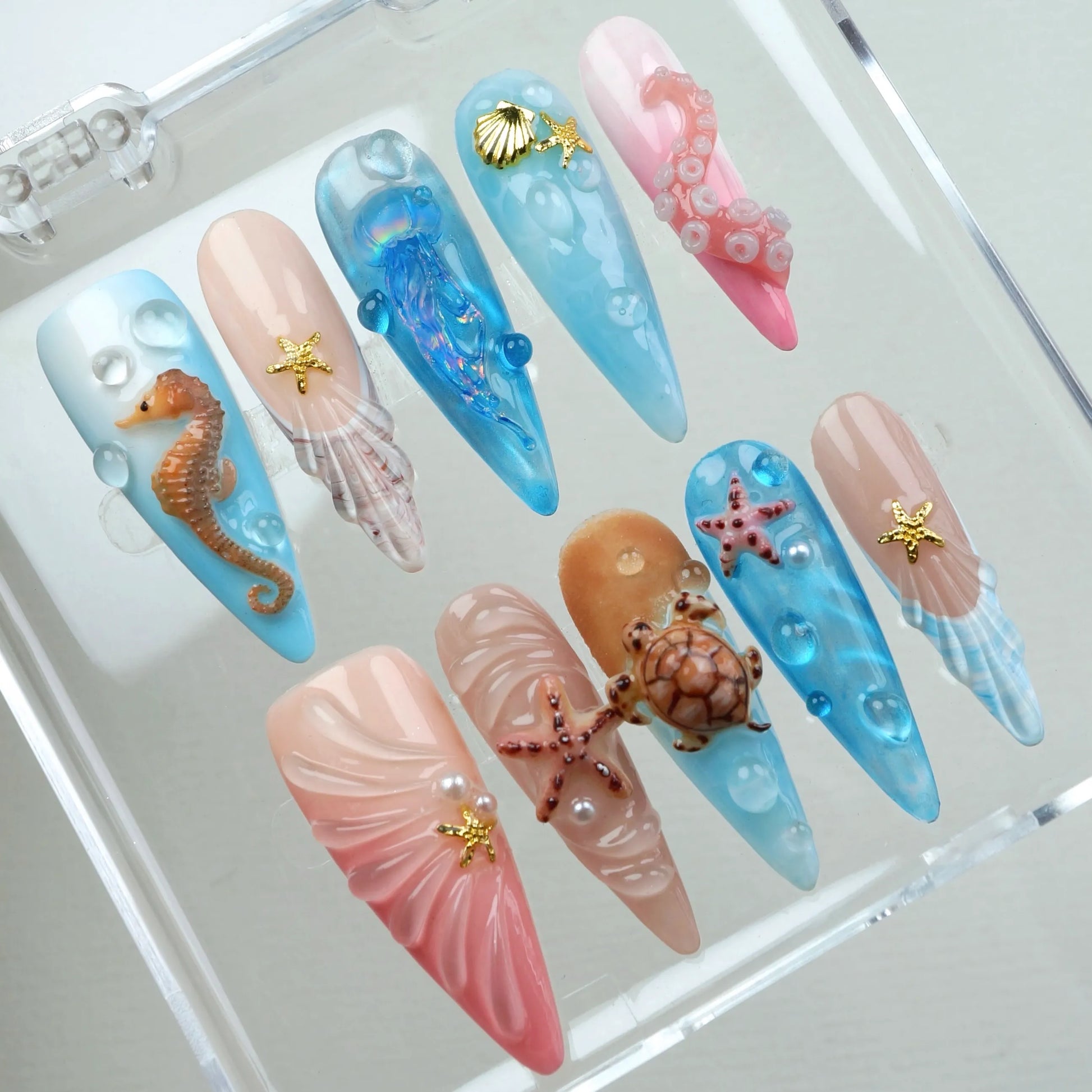 Aquatic press-on nail set in clear acrylic display case showing all 10 stiletto nails with 3D seahorse, sea turtle, jellyfish, and starfish ocean charms on baby blue, nude pink, coral, and sandy beige nail bases with water bubble art
