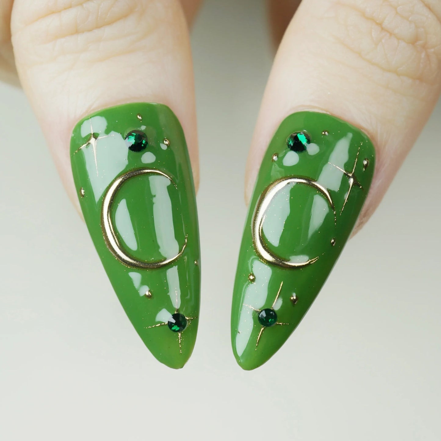 Green nail art with gold rings and small black stones on a neutral background