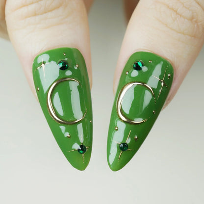 Green nail art with gold rings and small black stones on a neutral background