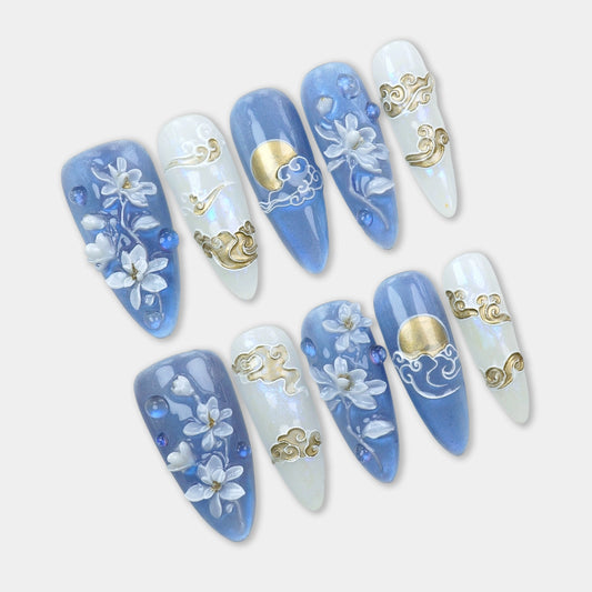 Press-on nails with hand-sculpted white florals, golden moon and cloud details, and soft blue and pearl shimmer base by Donailsart