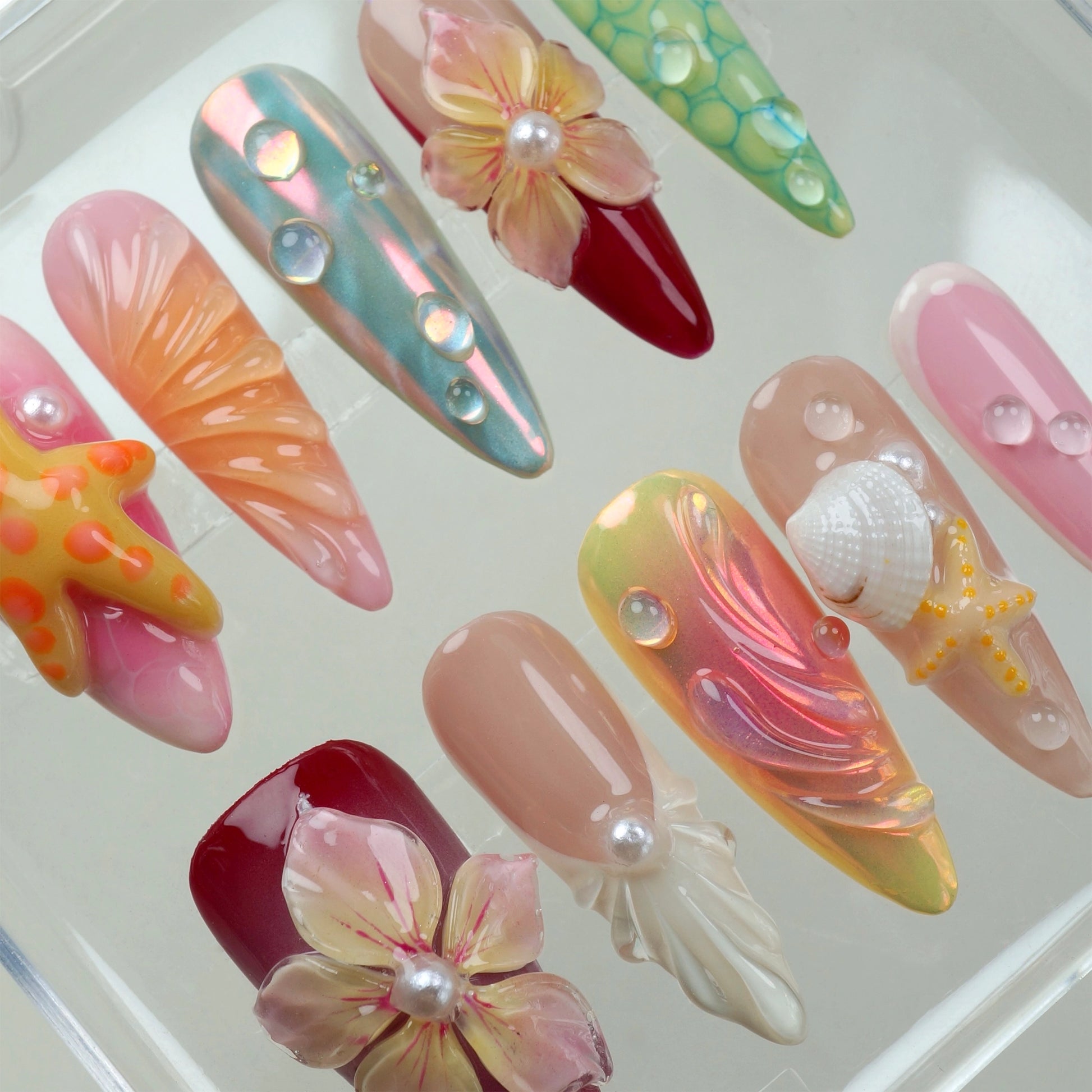 Handcrafted ocean-themed press-on nails with 3D starfish, seashells, and floral designs in pink and iridescent shades, displayed in a clear case
