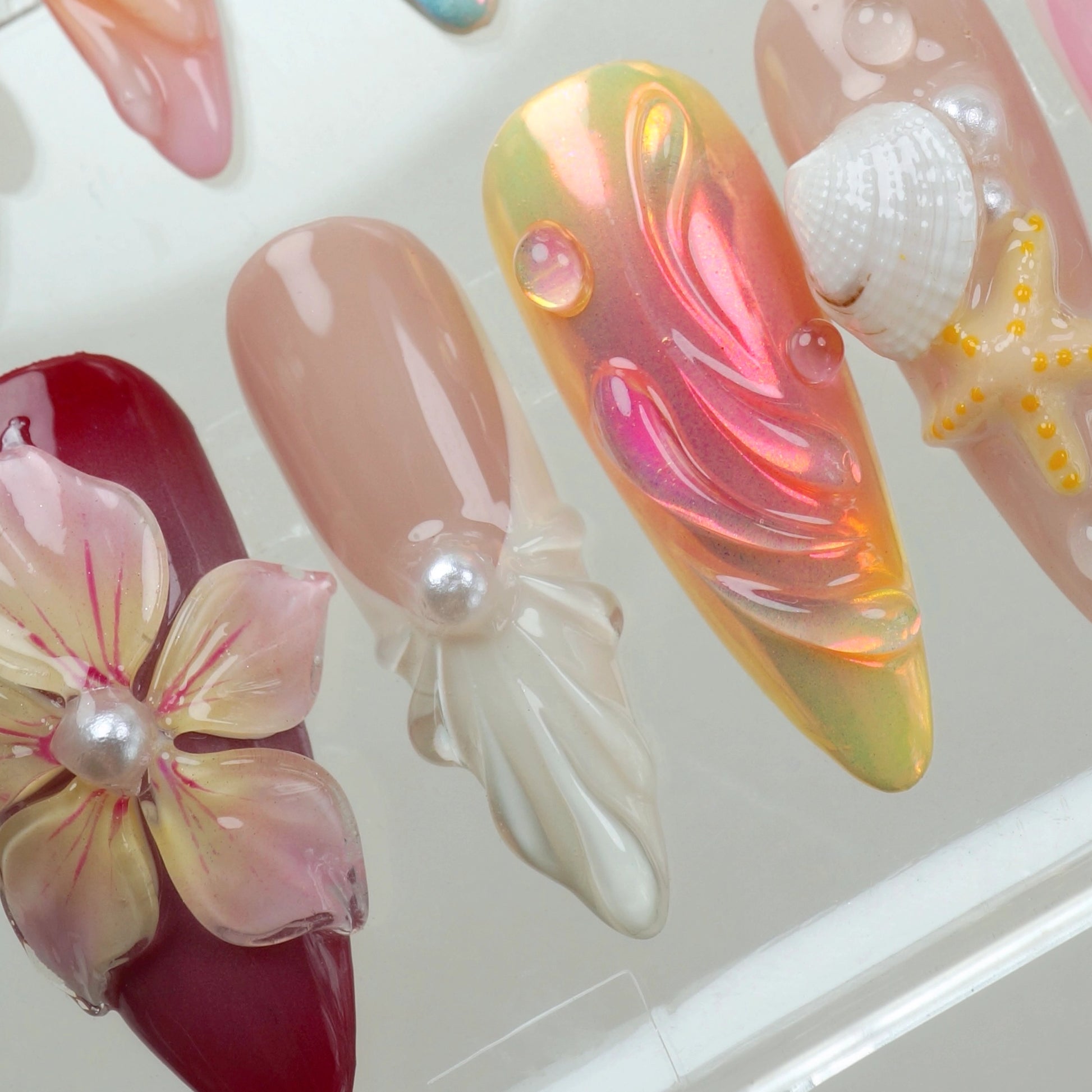 Handcrafted ocean-themed press-on nails with 3D starfish, seashells, and floral designs in pink and iridescent shades, displayed in a clear case