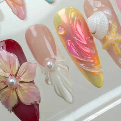 Handcrafted ocean-themed press-on nails with 3D starfish, seashells, and floral designs in pink and iridescent shades, displayed in a clear case
