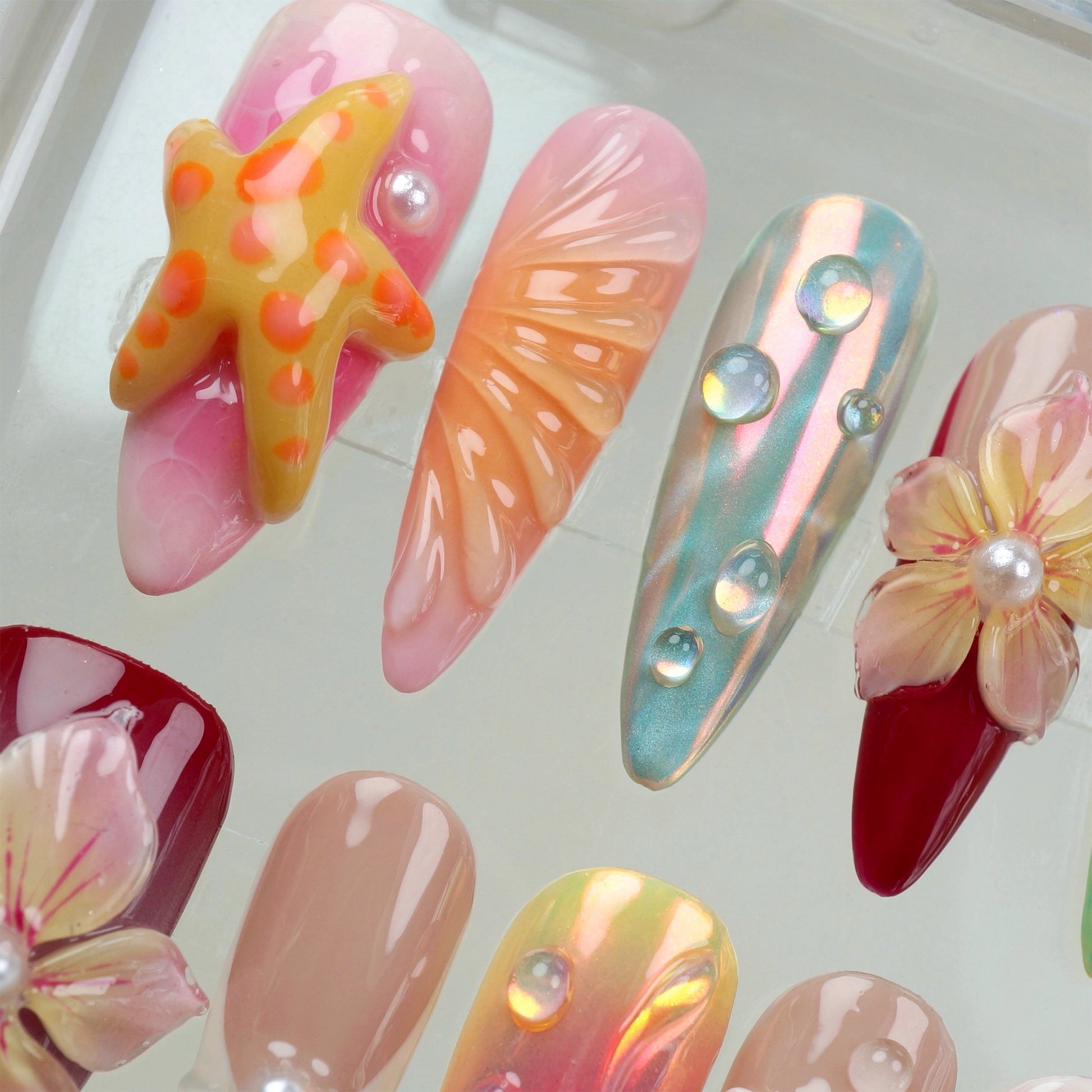 Handcrafted ocean-themed press-on nails with 3D starfish, seashells, and floral designs in pink and iridescent shades, displayed in a clear case