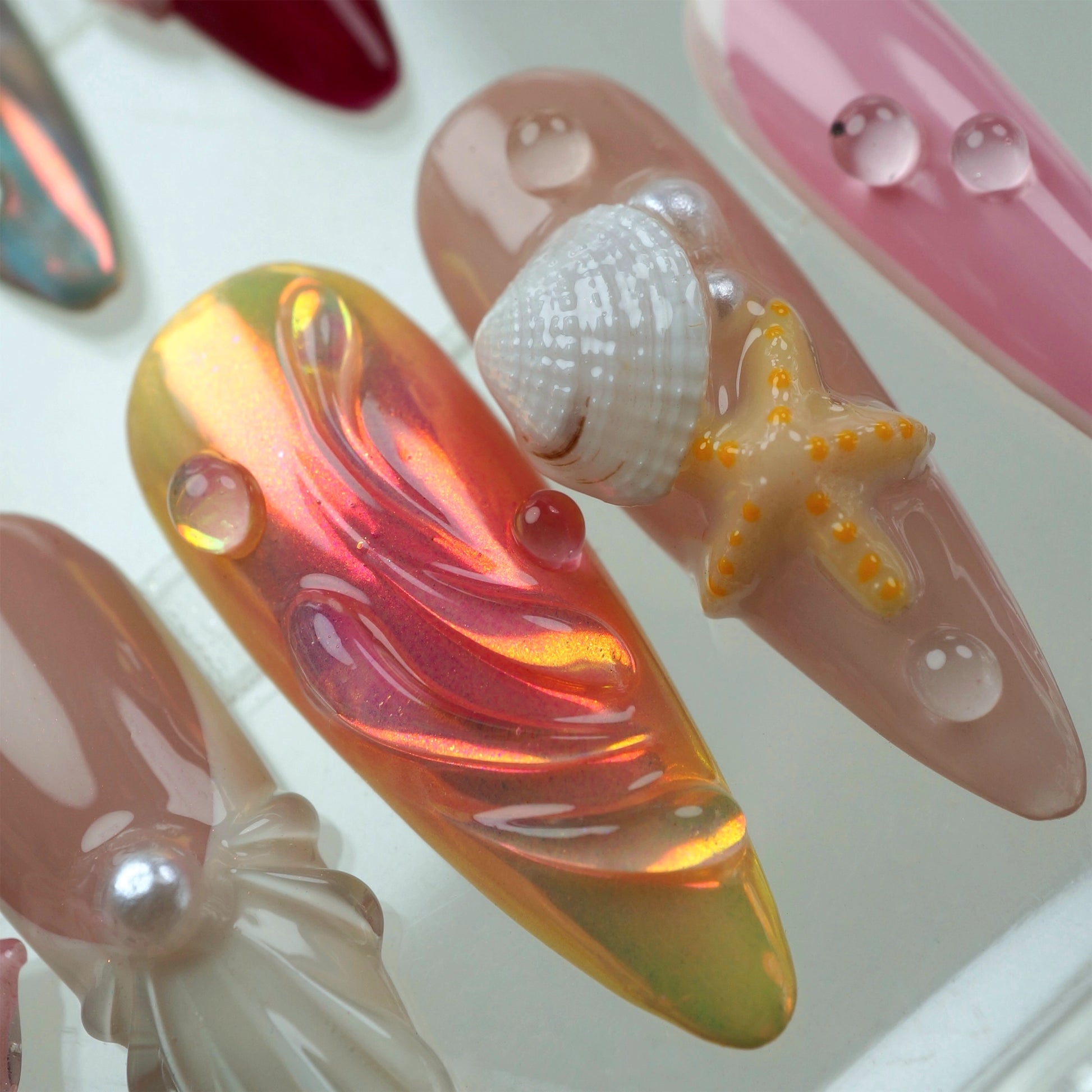 Handcrafted ocean-themed press-on nails with 3D starfish, seashells, and floral designs in pink and iridescent shades, displayed in a clear case