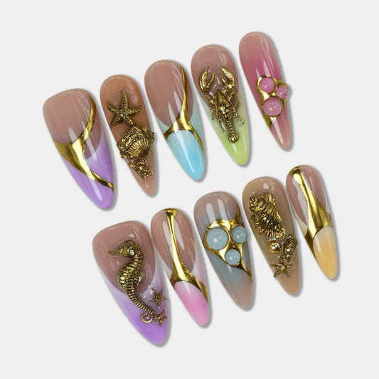 Ocean-inspired press-on nails from Donailsart with gold seashell charms, pastel ombré tips, and metallic swirl details