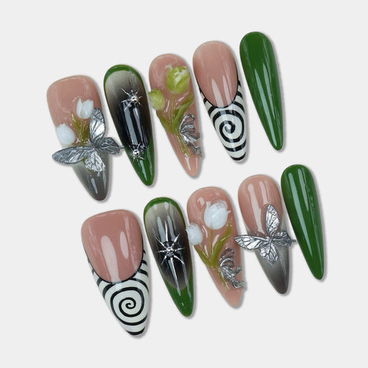 Elegant flower nails featuring 3D white floral accents, silver butterflies, green and black ombré gradients, and spiral patterns on nude-toned press-ons in a clear case