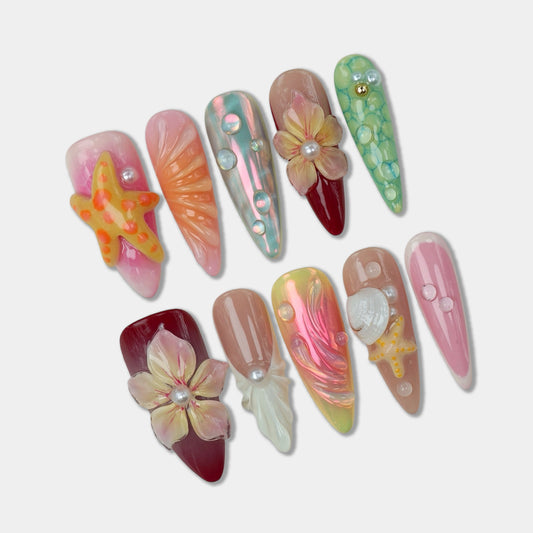Handcrafted ocean-themed press-on nails with 3D starfish, seashells, and floral designs in pink and iridescent shades, displayed in a clear case